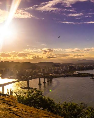 Pôr do sol visto do alto de uma pedra, com a luz dourada sobre a baía, a Terceira Ponte e a cidade ao fundo.
